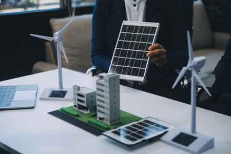 Two Young Engineers Expertise in Solar Cell Installation Meetings and Discussion in The Job. Planning to Install Solar Photovoltaic Panels on Roof Top in The Office Room with Factory Building Plan.の写真素材