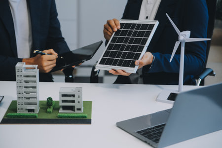 Two Young Engineers Expertise in Solar Cell Installation Meetings and Discussion in The Job. Planning to Install Solar Photovoltaic Panels on Roof Top in The Office Room with Factory Building Plan.の写真素材