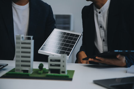 Two Young Engineers Expertise in Solar Cell Installation Meetings and Discussion in The Job. Planning to Install Solar Photovoltaic Panels on Roof Top in The Office Room with Factory Building Plan.の写真素材