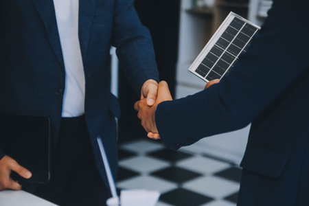 Two Young Engineers Expertise in Solar Cell Installation Meetings and Discussion in The Job. Planning to Install Solar Photovoltaic Panels on Roof Top in The Office Room with Factory Building Plan.の写真素材