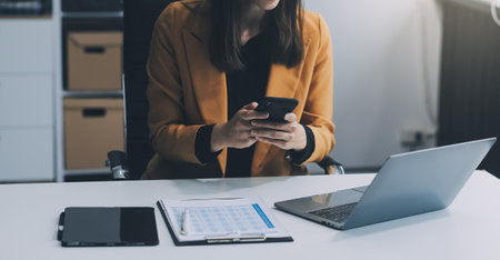 Beautiful Asian woman using a smartphone and working with a laptop while sitting at an office desk,...