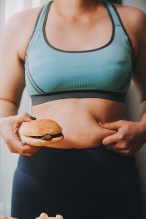 Portrait of obese woman looks sad while pinching her belly fat surrounded by junk foods. Diet fail conceptの写真素材