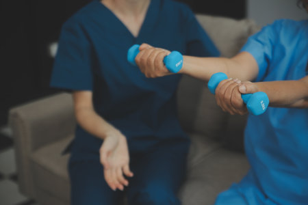 Old woman training with physiotherapist using dumbbells at home. Therapist assisting senior woman with exercises in nursing home. Elderly patient using dumbbells with outstretched arms.の写真素材