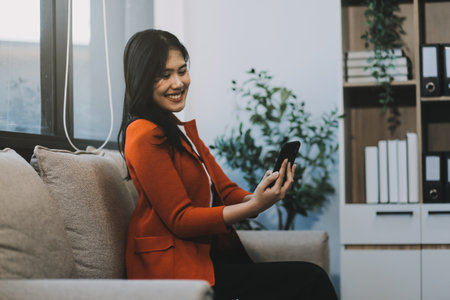 Smiling and happy millennial Asian businesswoman chatting with her friends on her phone, scrolling on social media or watching video on the internet, using her phone while relaxing in the office.の写真素材