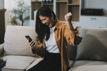 Emotional young asian woman in casual sitting on couch with laptop and raising hands up, happy...
