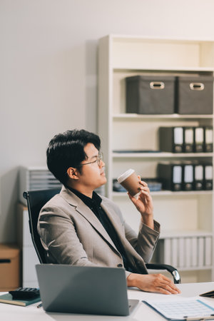 Asian businessmen wearing glasses Sipping coffee while sitting on the computer screen. Concept is to sit and work smart Happy Bright and Relaxed atmosphere. Background is a document rack.の写真素材