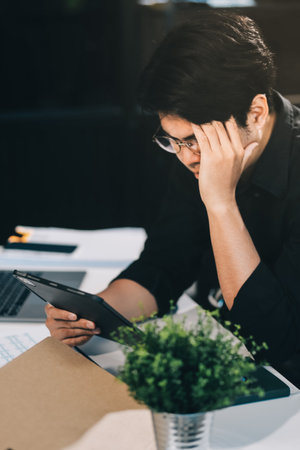 Frustrated Asian young businessman worker working on table in office. Busy employee worker people feel tired and headache from late work and overwork job document on computer desk at company workplaceの写真素材