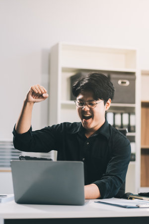 Celebrating success. Young asian business man working with laptop, tablet and papers on desk at office. He feeling good and happy.の写真素材