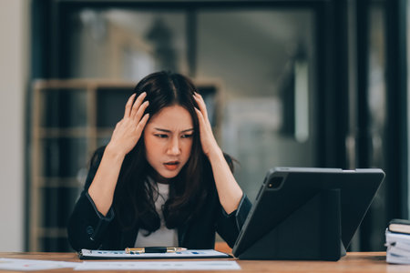 Young sad businesswoman is sitting at table, covering his face On desk is laptop, tablet computer, Stress. Serious woman concentrating on his paper work.の写真素材