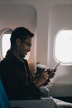 Happy curly man use phone and listens to music headphones on board airplane during flight.