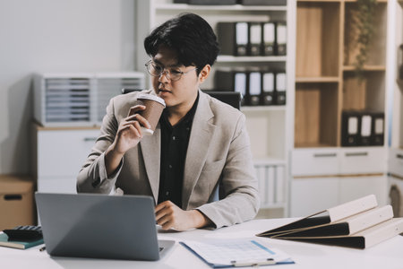 Asian businessmen wearing glasses Sipping coffee while sitting on the computer screen. Concept is to sit and work smart Happy Bright and Relaxed atmosphere. Background is a document rack.の写真素材