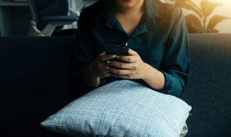 Charming young Asian woman using cellphone, communicating on web, working or learning online, sitting in armchair against white wall, free space. Lovely millennial lady chatting on smartphoneの写真素材