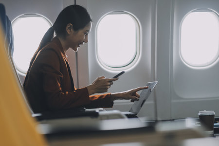 Asian female tourist checks notifications on smartphone while seated on airplane A businesswoman happily shares media from her phone, taking photos of the view from the window during a flight.の写真素材