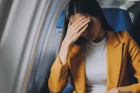 Young asian chinese airplane passenger woman experiencing discomfort, Airplane headache. Worried female passenger holding head, feeling unwell Anxiety and fear in the skies. Stressful travel moments.の写真素材