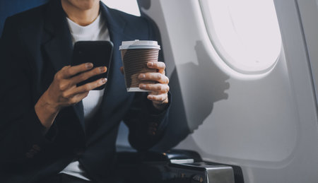 Young business woman sitting inside plane at the airport with sky view from the window drinking a...