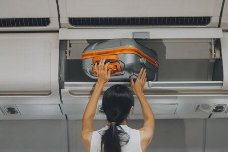 Young Female Tourist Storing Luggage in Overhead Compartment on Airplane During Travelの写真素材