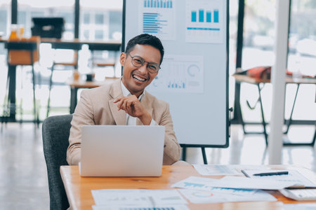 Excited asian male entrepreneur using smartphone and laptop, working online while sitting at workplace at home office. Businessman texting via cellphone and smilingの写真素材