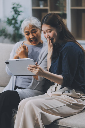 Happy grownup daughter showing content on tablet to mature mother. Two family generations women with digital computer gadget resting on couch together, watching videos, making video callの写真素材
