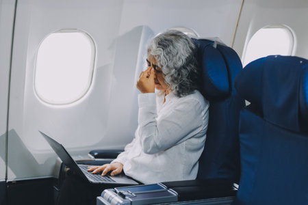 A mature woman sits by the window on a passenger plane, traveling abroad. She looks unwell, experiencing a headache, nausea, and vomiting, which dampens her travel excitement.の写真素材