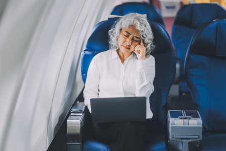 A mature woman sits by the window on a passenger plane, traveling abroad. She looks unwell, experiencing a headache, nausea, and vomiting, which dampens her travel excitement.の写真素材