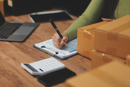 Startup SME small business entrepreneur of freelance Asian woman wearing apron using laptop and box to receive and review orders online to prepare to pack sell to customers, online sme business ideas.の写真素材