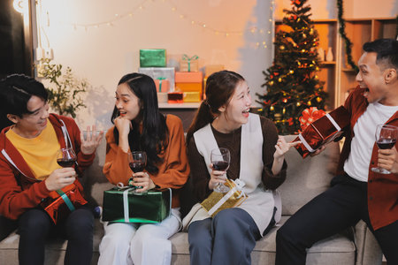 Group of young Asian man and women as friends having fun at a New Year's celebration, holding gift boxes standing by Christmas tree decoration, midnight countdown Party at home with holiday season.の写真素材