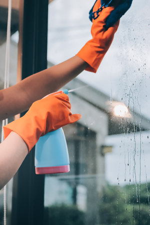 Young female worker cleaning window glass with cloth in spacious villa