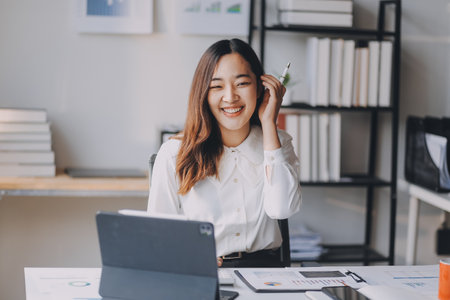 Young asian businesswoman sitting on an ergonomic chair and drinking coffee while working on a laptop computer in a modern office, she is smiling and looking at the screenの写真素材