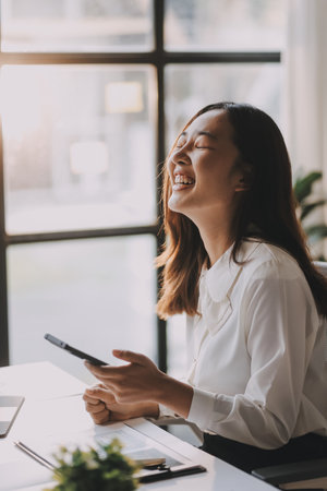 Young asian woman talking on mobile phone while taking notes and working with laptop computer at home. Business, home office and technology concept.の写真素材
