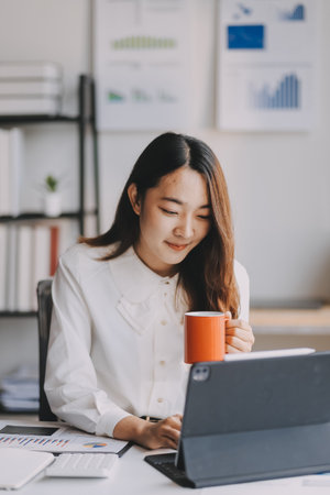 Young asian businesswoman sitting on an ergonomic chair and drinking coffee while working on a laptop computer in a modern office, she is smiling and looking at the screenの写真素材