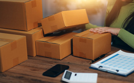 Startup SME small business entrepreneur of freelance Asian woman wearing apron using laptop and box to receive and review orders online to prepare to pack sell to customers, online sme business ideas.の写真素材