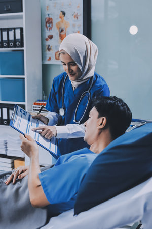 A young man lies on a bed in a hospital ward talking with her young Indian doctor. Doctor checking patient's health doctor treating patient, hospital admission of patientの写真素材