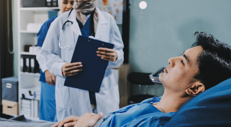 A young man lies on a bed in a hospital ward talking with his young doctor. Doctor checking patient's health, treating patient, hospital admission of patientの写真素材