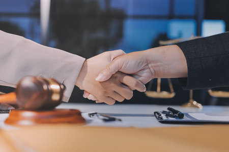 Two businesspeople are shaking hands in a law office with a scale and a gavel on the tableの写真素材