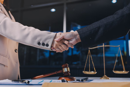 Two businesspeople are shaking hands in a law office with a scale and a gavel on the tableの写真素材