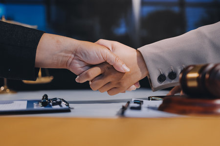 Two businesspeople are shaking hands in a law office with a scale and a gavel on the tableの写真素材