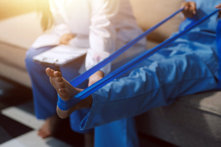 Confident physical therapist helps patient use resistance band stretching out his leg in clinic room.の写真素材