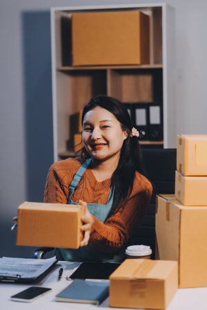 Startup SME small business entrepreneur of freelance Asian woman wearing apron using laptop and box to receive and review orders online to prepare to pack sell to customers, online sme business ideas.の写真素材