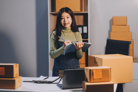 Startup SME small business entrepreneur of freelance Asian woman wearing apron using laptop and box to receive and review orders online to prepare to pack sell to customers, online sme business ideas.の写真素材