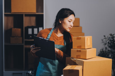 Startup SME small business entrepreneur of freelance Asian woman wearing apron using laptop and box to receive and review orders online to prepare to pack sell to customers, online sme business ideas.の写真素材