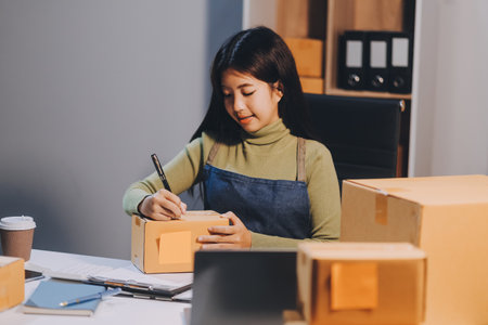 Startup SME small business entrepreneur of freelance Asian woman wearing apron using laptop and box to receive and review orders online to prepare to pack sell to customers, online sme business ideas.の写真素材