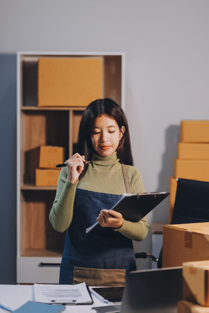 Startup SME small business entrepreneur of freelance Asian woman wearing apron using laptop and box to receive and review orders online to prepare to pack sell to customers, online sme business ideas.の写真素材