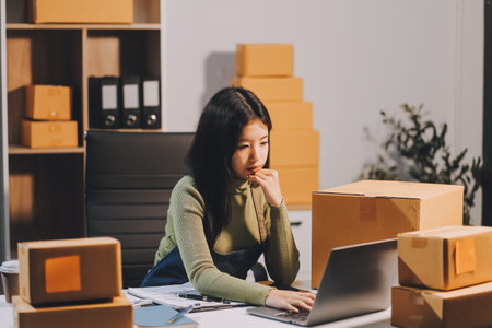 Startup SME small business entrepreneur of freelance Asian woman wearing apron using laptop and box to receive and review orders online to prepare to pack sell to customers, online sme business ideas.の写真素材