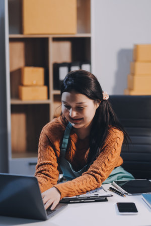 Startup SME small business entrepreneur of freelance Asian woman wearing apron using laptop and box to receive and review orders online to prepare to pack sell to customers, online sme business ideas.の写真素材