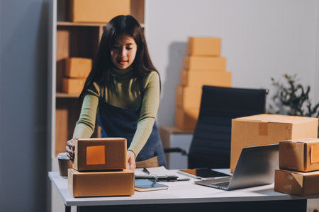 Startup SME small business entrepreneur of freelance Asian woman wearing apron using laptop and box to receive and review orders online to prepare to pack sell to customers, online sme business ideas.の写真素材