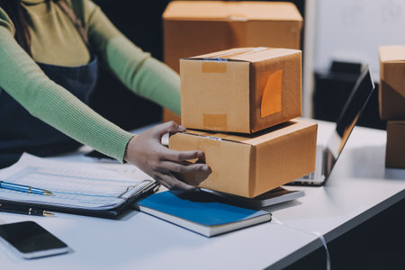 Startup SME small business entrepreneur of freelance Asian woman wearing apron using laptop and box to receive and review orders online to prepare to pack sell to customers, online sme business ideas.の写真素材