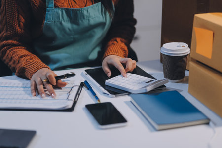 Startup SME small business entrepreneur of freelance Asian woman wearing apron using laptop and box to receive and review orders online to prepare to pack sell to customers, online sme business ideas.の写真素材