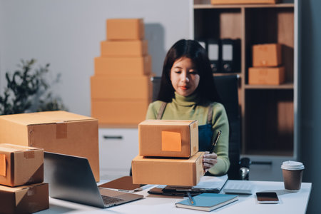 Startup SME small business entrepreneur of freelance Asian woman wearing apron using laptop and box to receive and review orders online to prepare to pack sell to customers, online sme business ideas.の写真素材