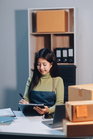 Startup SME small business entrepreneur of freelance Asian woman wearing apron using laptop and box to receive and review orders online to prepare to pack sell to customers, online sme business ideas.の写真素材