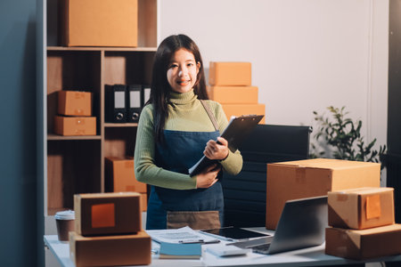 Startup SME small business entrepreneur of freelance Asian woman wearing apron using laptop and box to receive and review orders online to prepare to pack sell to customers, online sme business ideas.の写真素材
